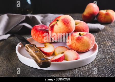 Whole and sliced peaches and a knife are placed on a plate on the table. Stock Photo