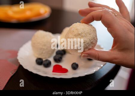 Close up of a female hand reaching for a white coconut truffle candy on a plate decorated with fresh blueberries and a small red heart. Concept of swe Stock Photo