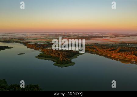Aerial sunrise over Masurian Lakeland in Poland, mist drifting above calm lakes and patchwork fields, golden morning light illuminating forests and ru Stock Photo