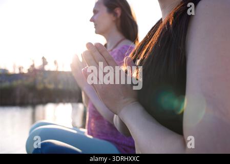 Two women practicing mindfulness meditation by the water, hands in prayer position, with sunlight illuminating their serene expr Stock Photo