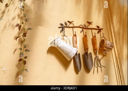Vintage gardening tools like trowels and a small watering can hanging on a yellow wall. Concept of home gardening hobby, domestic plant care, and rust Stock Photo