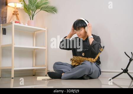 Young Asian man playing saxophone indoors in a cozy room with soft lighting Stock Photo