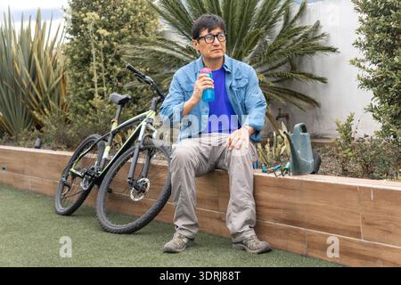 Asian man sitting outdoors drinking from a bottle, resting next to a bicycle Stock Photo