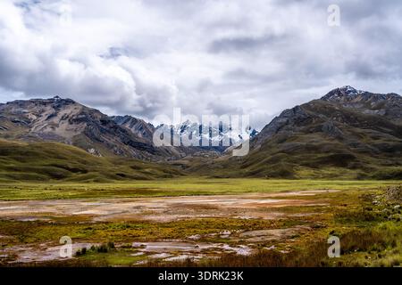 A marshy lake reflecting the dramatic cloudy sky is surrounded by rolling hills and snow-capped peaks on the Pastoruri Glacier t Stock Photo
