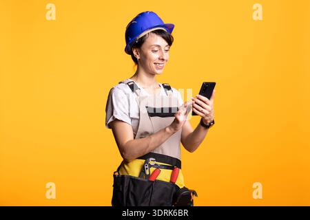 Caucasian female builder in safety helmet cheerfully using cellphone, reading funny message or instruction. Woman contractor stands with tool belt, reflecting onsite coordination and mobile updates. Stock Photo