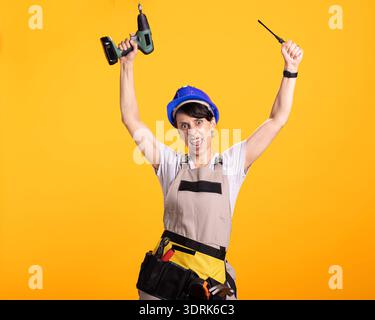 Happy white woman in overalls and tool belt lifts drill gun and screwdriver with excitement. Caucasian female builder dances and sings in studio, having fun with construction tools. Stock Photo