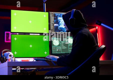 Hacker listening music doing covert operations, using green screen PC to bypass security. Rogue engineer wearing headphones engineering data theft script to penetrate firewalls using mockup computer Stock Photo