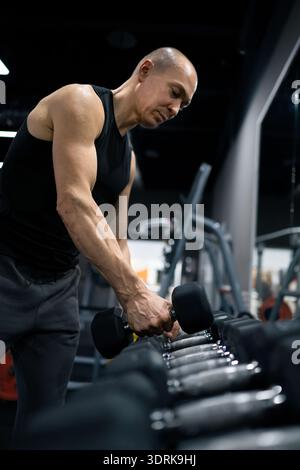 Muscular man holding dumbbell and resting after strength training session in modern fitness gym. Recovery and determination concept. Stock Photo