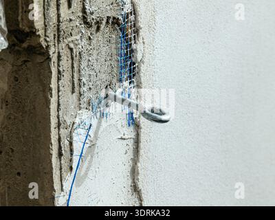 A close-up of a construction detail showing a metal hook set into a freshly plastered wall, with blue reinforcement mesh visible beneath the surface. Stock Photo