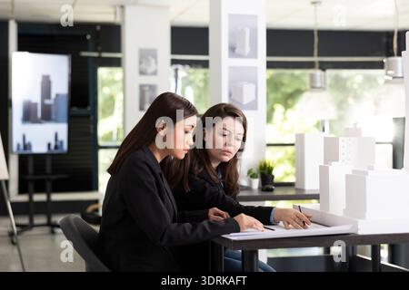 Female architects review blueprint details and discuss layout ideas in modern design studio. Dedicated diverse professionals examine architectural models, assessing measurements and design accuracy. Stock Photo
