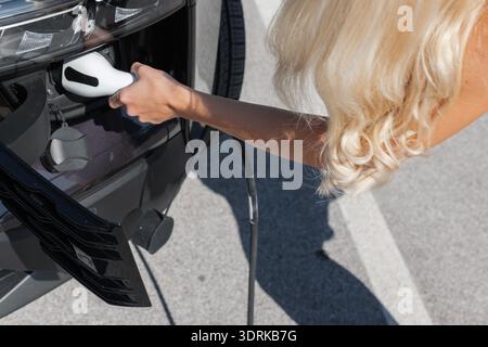 A woman inserts a charging plug into an electric car at a charging station during daylight in a parking lot. Stock Photo