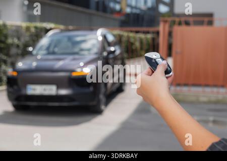 Woman stands holding a car key remote unlocking her parked car in an urban setting during daytime. Stock Photo
