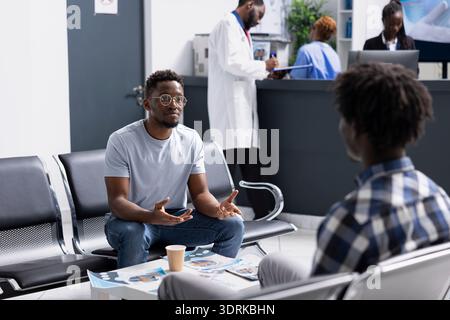 Two male clients sit together in hospital waiting room, speaking before scheduled medical checkups. African American patient preparing for doctor appointment while waiting in clinical lobby. Stock Photo