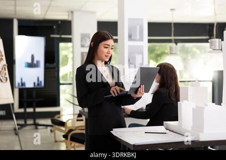 Young female architect uses laptop while analyzing 3d building models. Focused professional reviews project plans, combining digital tools with physical designs for accurate architectural development. Stock Photo