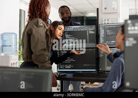 African American and Asian colleagues work side by side, analyzing source code and data on desktop monitors. Focused programmers collaborate to enhance web development projects in modern office. Stock Photo