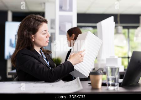 Young architect carefully reviews floor plans and 3D structure models, ensuring accurate measurements and project requirements. Busy employee prepares reports for ongoing construction development. Stock Photo