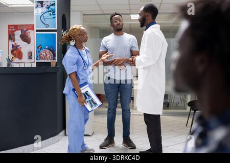 Doctor and nurse speaking with patient, addressing medical concerns in hospital lobby. Black man reviewing treatment plan with professional healthcare team in clinic reception area. Stock Photo