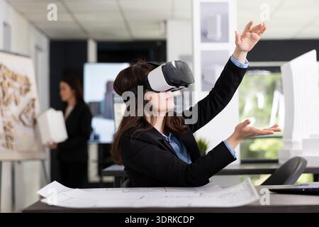 Young woman wears VR headset, reviewing building plans on laptop inside modern office. Female architect examines 3D layouts and floor plans as part of design and planning workflow with immersive tech. Stock Photo