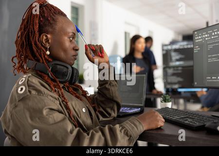 African american woman programmer deep in thought at her workstation, pen at temple, surrounded by multiple screens and colleagues in modern collaborative tech office workspace. Stock Photo