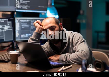 Tired programmer naps at workstation after extended hours handling cybersecurity issues. Bearded man sleeps on arm, overwhelmed by debugging tasks and mental strain of working with encrypted programs. Stock Photo