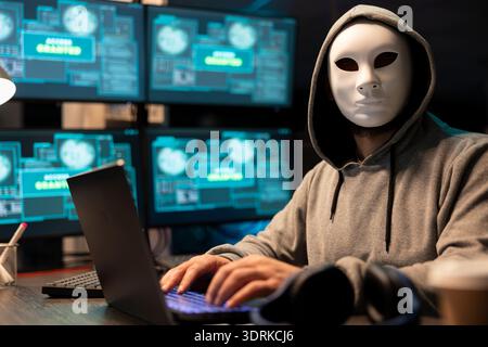 Anonymous man with white mask types on laptop surrounded by multiple monitors displaying system access visuals. Portrait of hacker at desk, attempting security breach on private company software. Stock Photo