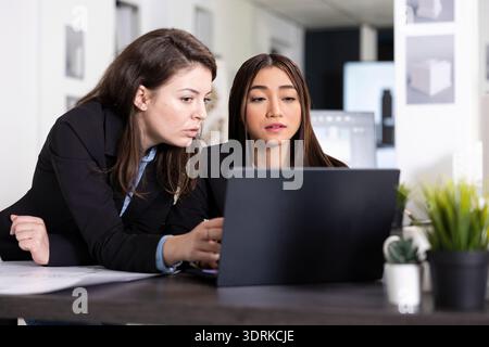 Focused women collaborate at desk, reviewing blueprints and planning architectural layout with precision. Female employees draft designs, preparing construction project reports for client presentation Stock Photo