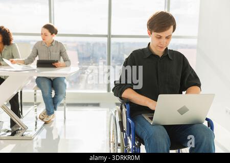Accessible environment. Young man in chair for people with disability working with laptop in modern office. Diverse multiracial Stock Photo