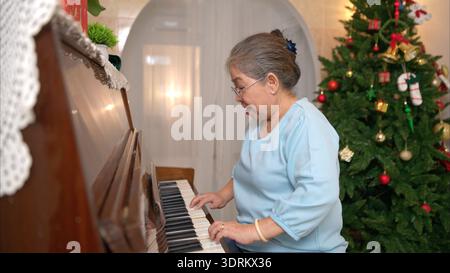 On a holiday, an elderly woman enjoys decoration Christmas tree and playing the piano at home Stock Photo