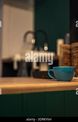 A teal ceramic coffee cup sits on a wooden countertop in a modern kitchen. A chrome faucet, white sink, and wooden spice box appear soft behind deep g Stock Photo