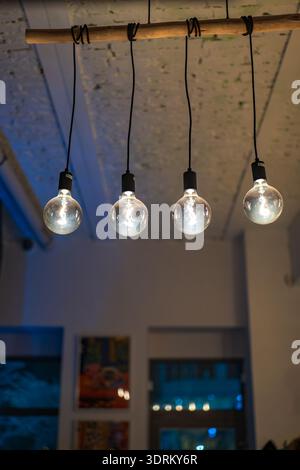 Four round bulbs hang from black cords on a rustic wood rod in a cafe or studio. Warm light glows, a blue window suggests evening, and blur isolates t Stock Photo
