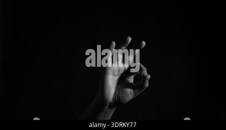 Low key monochrome close-up of hand making OK symbol, textured skin detail under directional light, isolated on dark background. Stock Photo