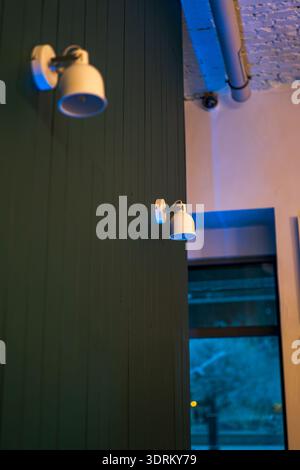 Two white sconces on a dark paneled partition emit cool light. A large window shows evening bokeh. An exposed brick ceiling with duct and a camera add Stock Photo