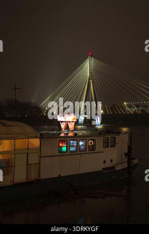 A moored riverboat with warm windows sits before Warsaw's Swietokrzyski Bridge, its single pylon and red beacon glow, fog softens skies, ripples light Stock Photo