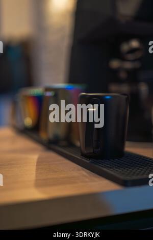 A matte black mug sits in sharp focus on a rubber bar mat, with rainbow and steel mugs behind, warm ambient light, bokeh, side lighting, evening cafe Stock Photo