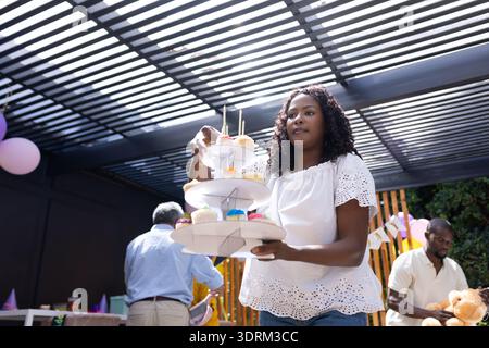 African American woman arranging colorful cupcakes on tiered stand at family party. Celebration, desserts, guest, gathering, pas Stock Photo