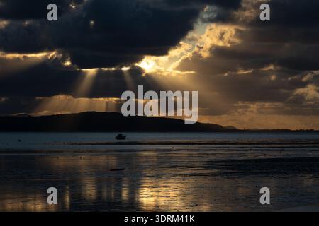 Golden sunlight rays breaking through dark clouds over a lone boat on the Dee Estuary Stock Photo