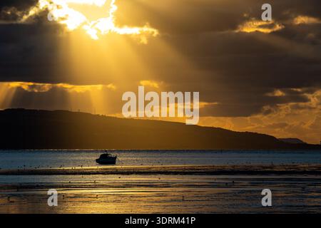 Golden sunlight rays breaking through dark clouds over a lone boat on the Dee Estuary Stock Photo
