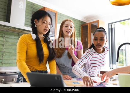 Teen girls collaborating on school project in kitchen, using tablet and papers. Collaboration, teamwork, brainstorming, technolo Stock Photo
