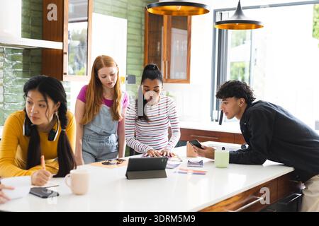 Teens collaborating on school project using tablet and smartphones in modern kitchen. Technology, teamwork, collaboration, educa Stock Photo