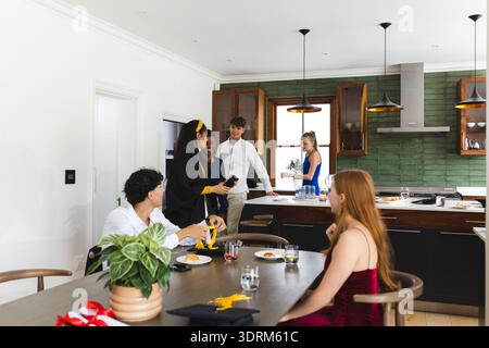 Celebrating graduation in kitchen, teens wearing caps and gowns, talking happily. celebration, achievement, friendship, teenager Stock Photo