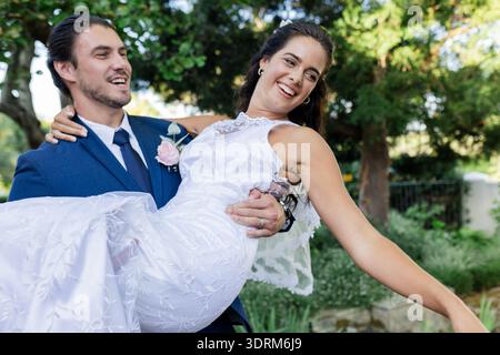 Groom joyfully carrying bride in garden, both smiling and celebrating their wedding. celebration, couple, marriage, happiness, l Stock Photo
