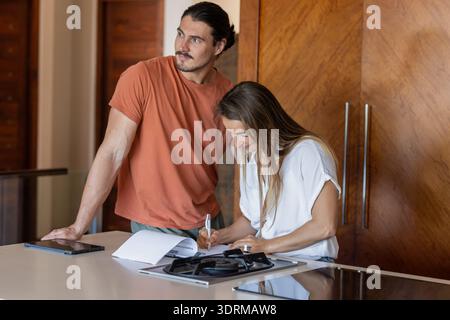 Couple in kitchen reviewing documents and using tablet for planning. Lifestyle, togetherness, teamwork, technology, home, collab Stock Photo