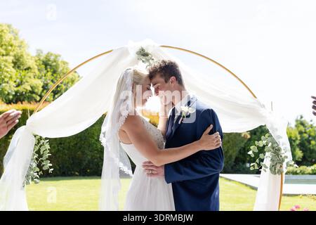 Bride and groom embracing under floral arch, sharing joyful moment outdoors. Wedding, couple, love, romance, celebration, embrac Stock Photo