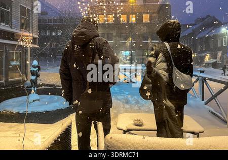 Two people in winter coats walking through heavy snowfall in Tromso, Norway at night, illuminated by warm city lights. Stock Photo