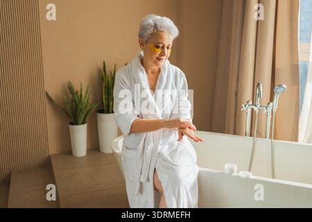Mature adult old woman applying moisturizing body lotion on hands. Senior lady rubbing nourishing anti age cream doing morning d Stock Photo