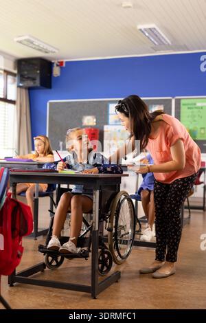 Asian teacher assists a girl in a wheelchair at a school classroom desk. Children are focused on their work in the background, i Stock Photo