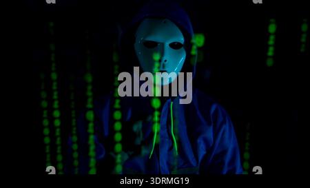 Anonymous hacker wearing a mask and hoodie interacting with digital data in a dark room Stock Photo