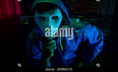 Anonymous hacker wearing a mask and hoodie interacting with digital data in a dark room Stock Photo