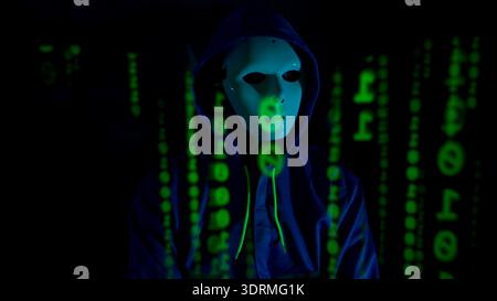 Anonymous hacker wearing a mask and hoodie interacting with digital data in a dark room Stock Photo