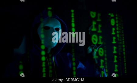 Anonymous hacker wearing a mask and hoodie interacting with digital data in a dark room Stock Photo